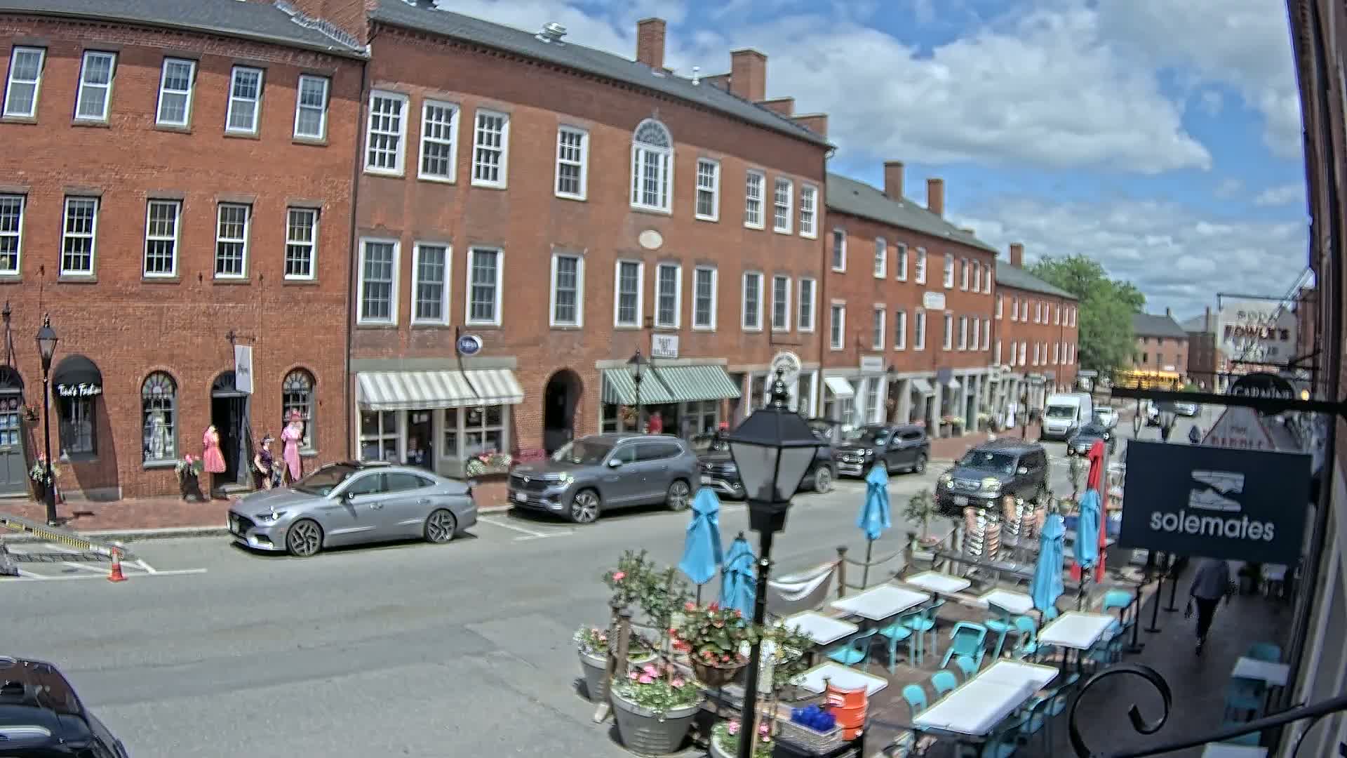 A sunny day on a brick-lined street shows several parked cars, pedestrians, and an outdoor restaurant patio with blue umbrellas and teal chairs.