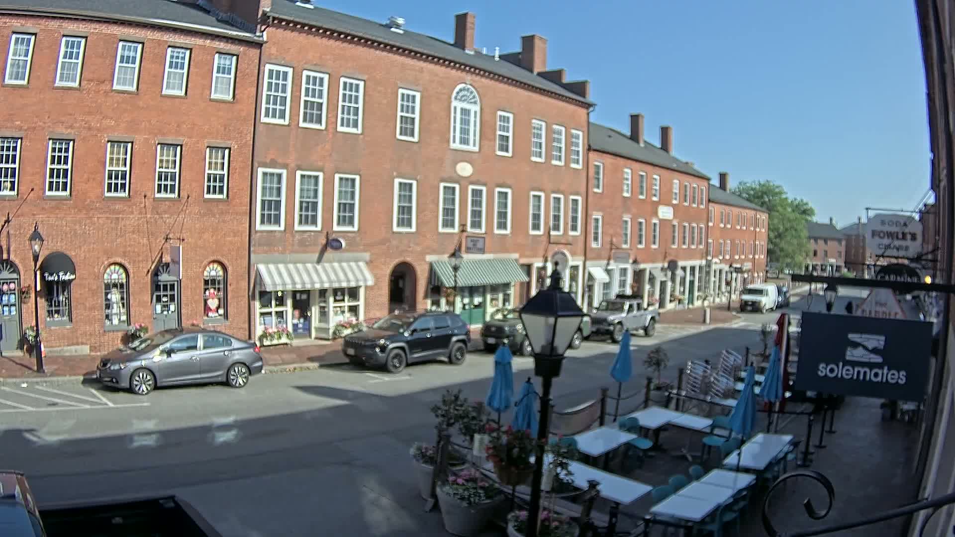 A sunny day reveals a brick street lined with shops and restaurants, outdoor seating with umbrellas, and several cars parked along the curb.