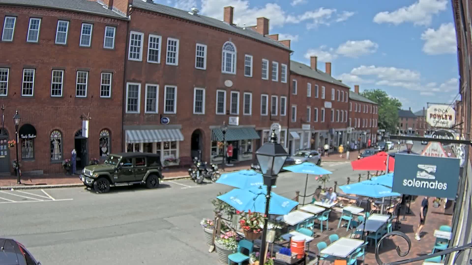 A sunny day on a brick-lined street with outdoor seating at a cafe in front of several shops and buildings.