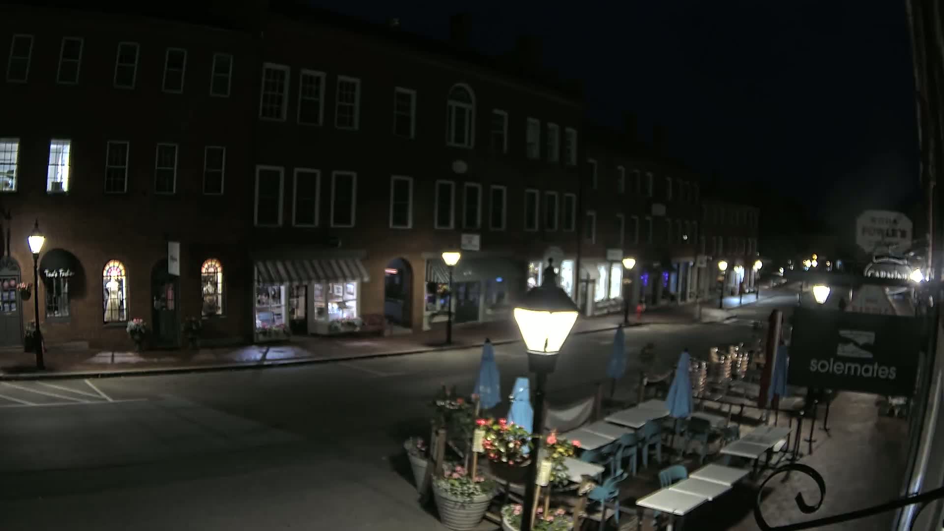 A nighttime street scene shows a row of dark brick buildings with illuminated storefronts and outdoor patio seating in front of a restaurant.