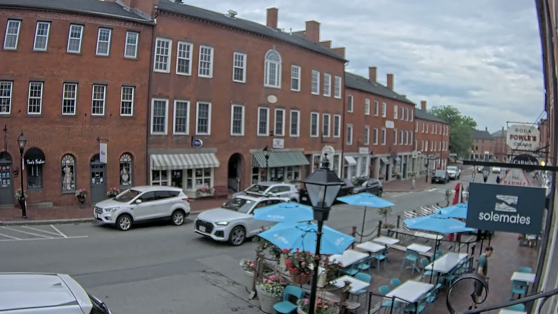 A street scene on an overcast day shows brick buildings with shops and restaurants, including an outdoor patio with light blue umbrellas and teal chairs.