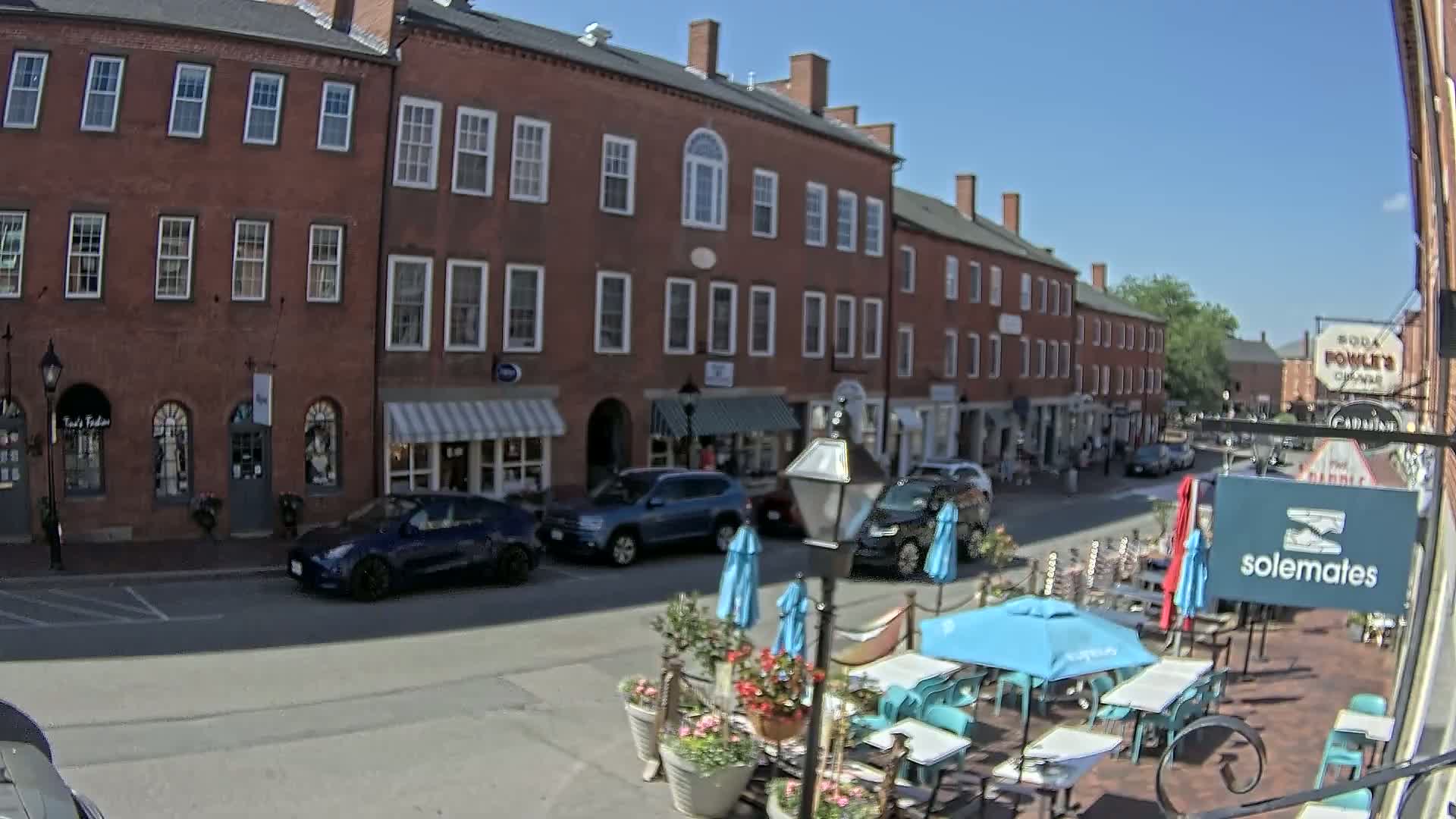 A sunny day shows a brick street lined with shops and restaurants, including outdoor seating with umbrellas.