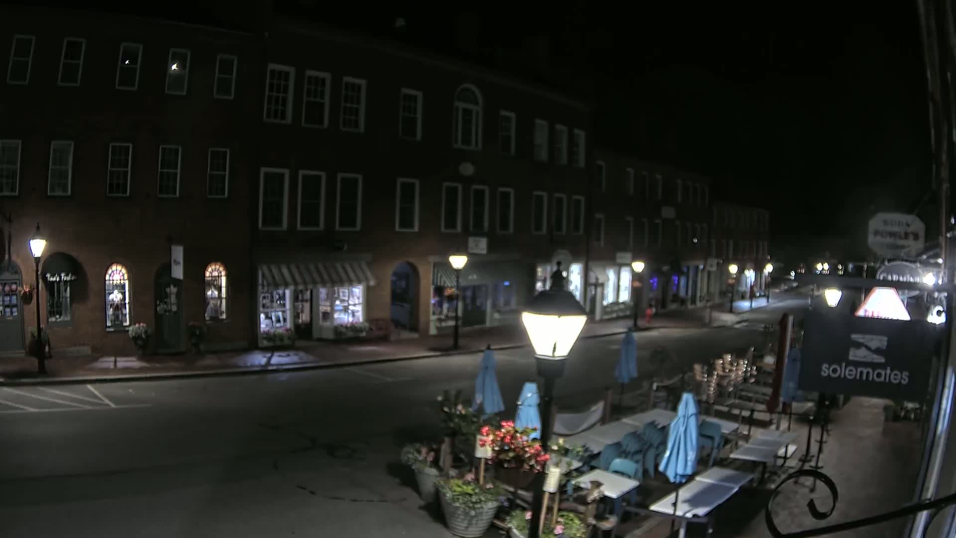 A nighttime street scene shows a row of dark brick buildings with outdoor seating and umbrellas in front of a restaurant.