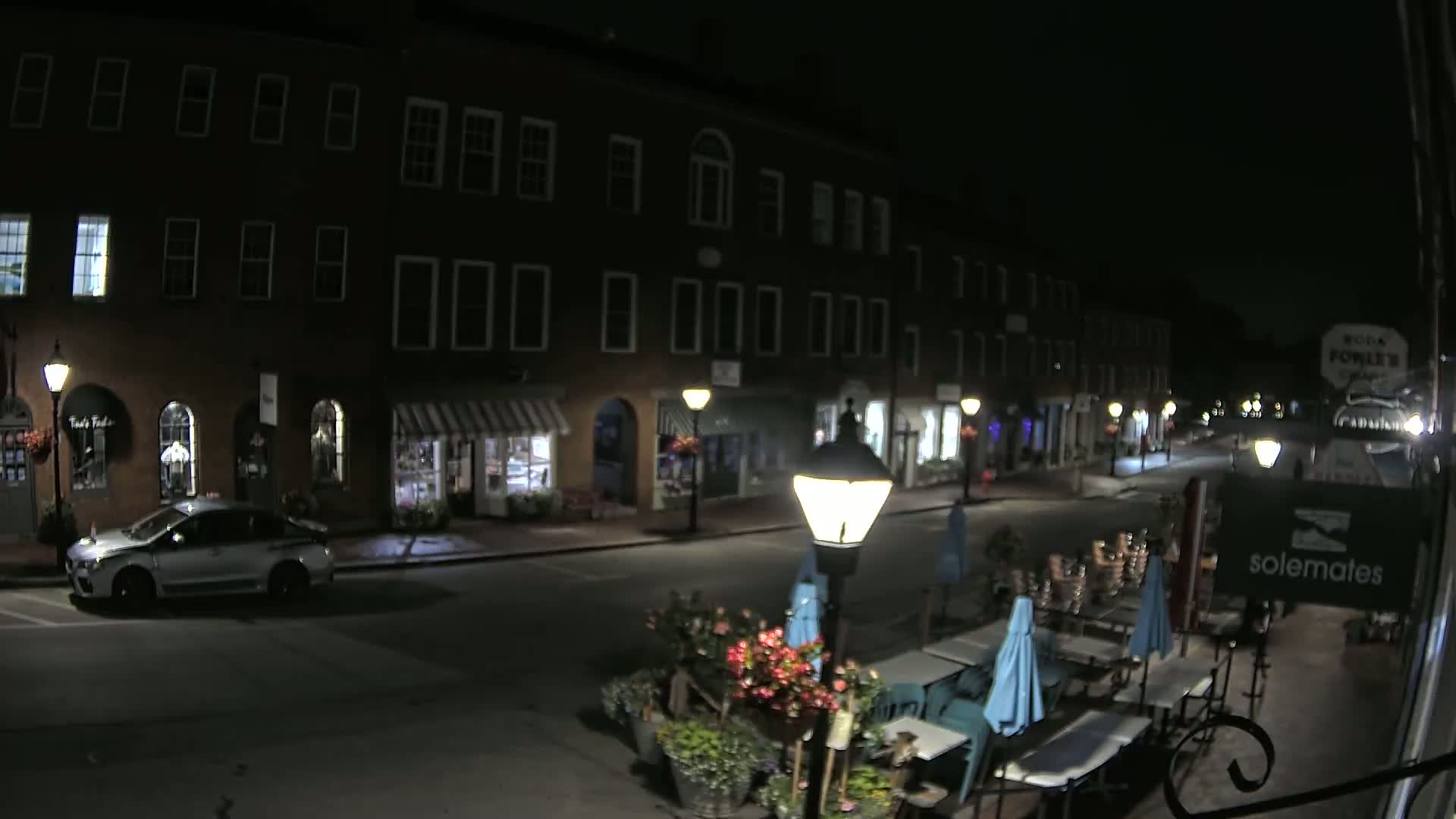 A nighttime street scene shows a row of dark brick buildings with shops and a parked car, with outdoor seating at a restaurant in the foreground under a clear sky.