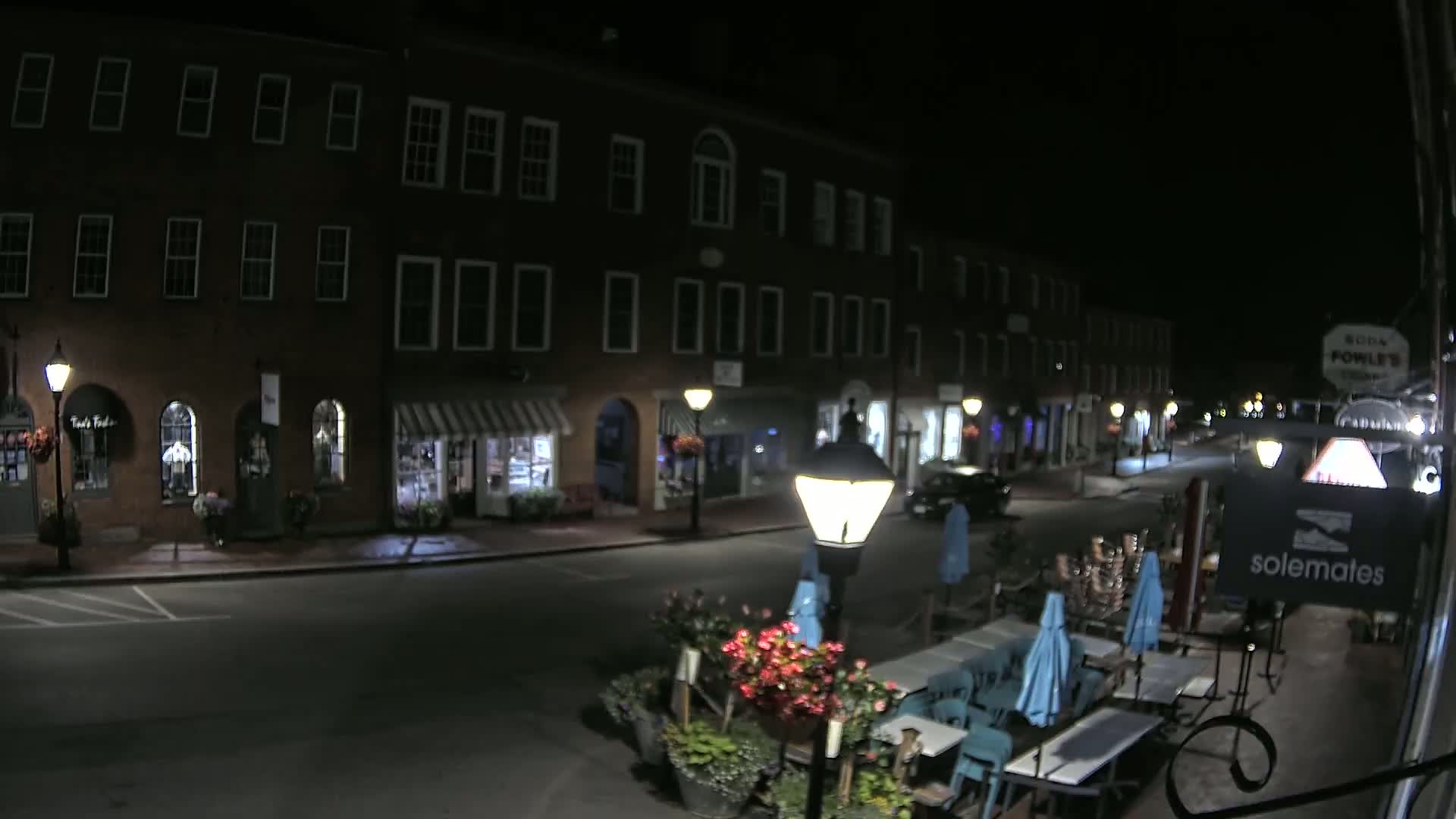 A nighttime street scene shows a row of brick buildings with outdoor seating at a restaurant in the foreground.
