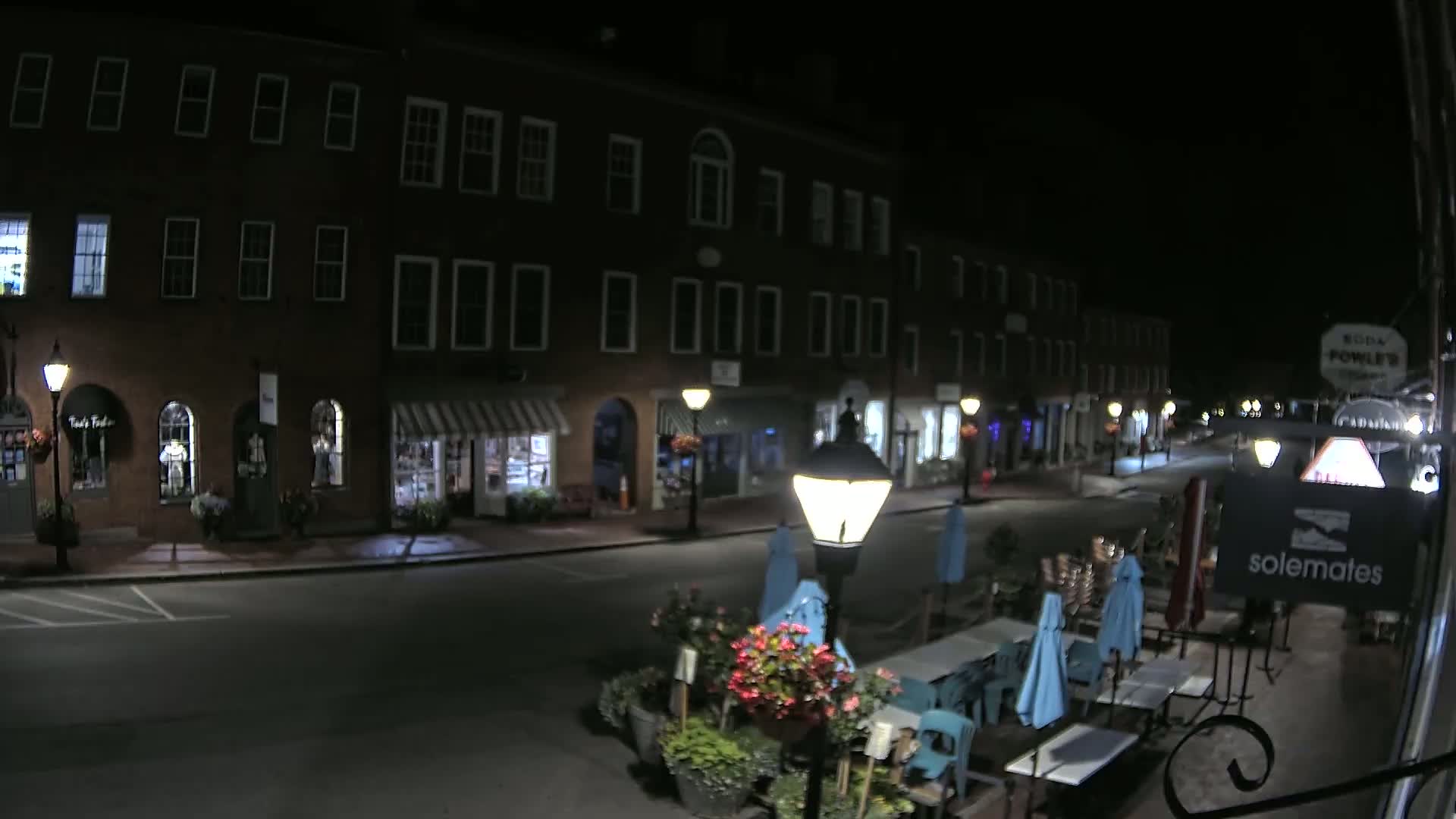 A nighttime street scene shows a row of brick buildings with shops and outdoor seating at a restaurant under a clear, dark sky.