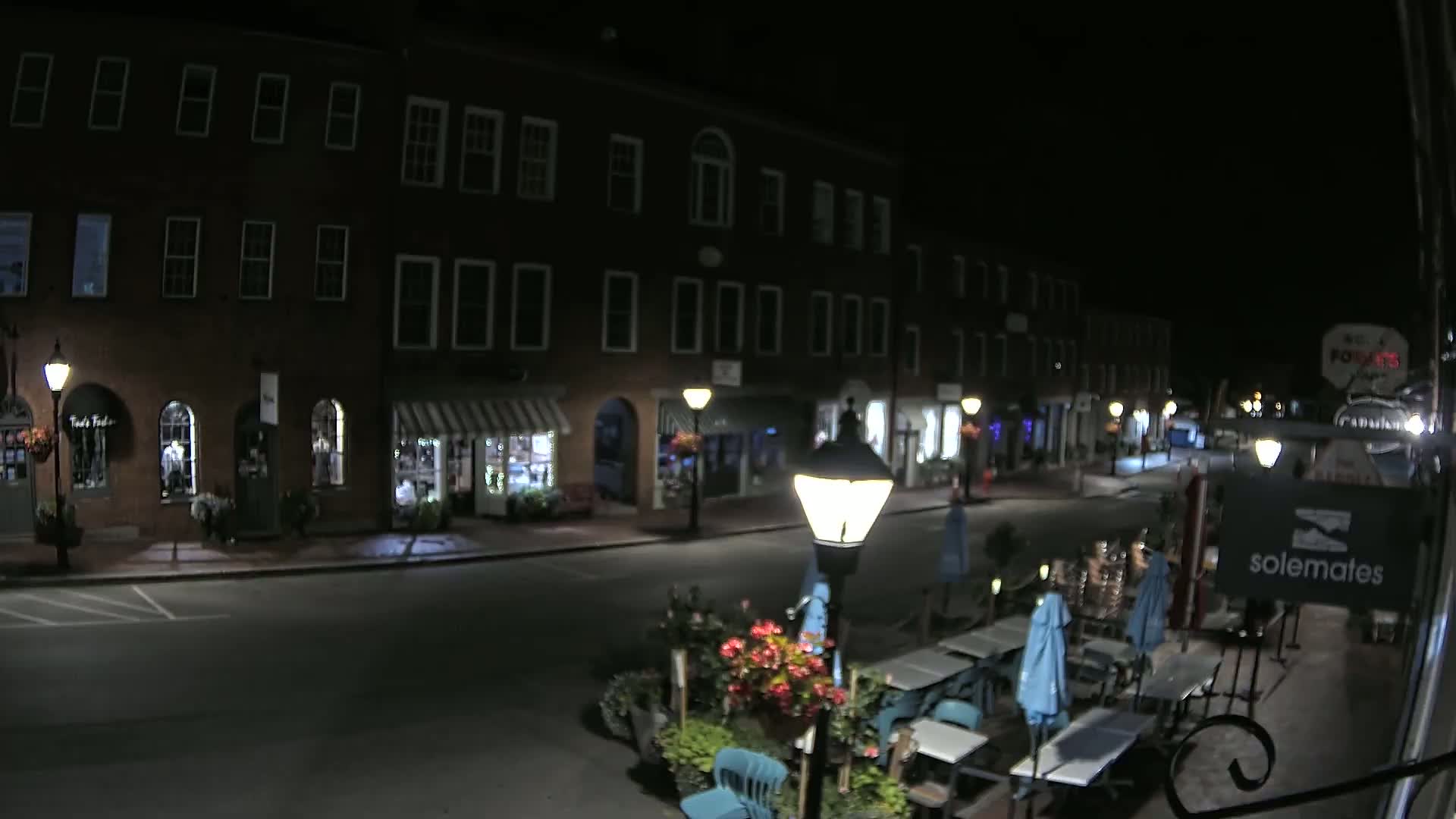 A nighttime street scene shows a row of brick buildings with shops and outdoor seating at a restaurant under a clear, dark sky.