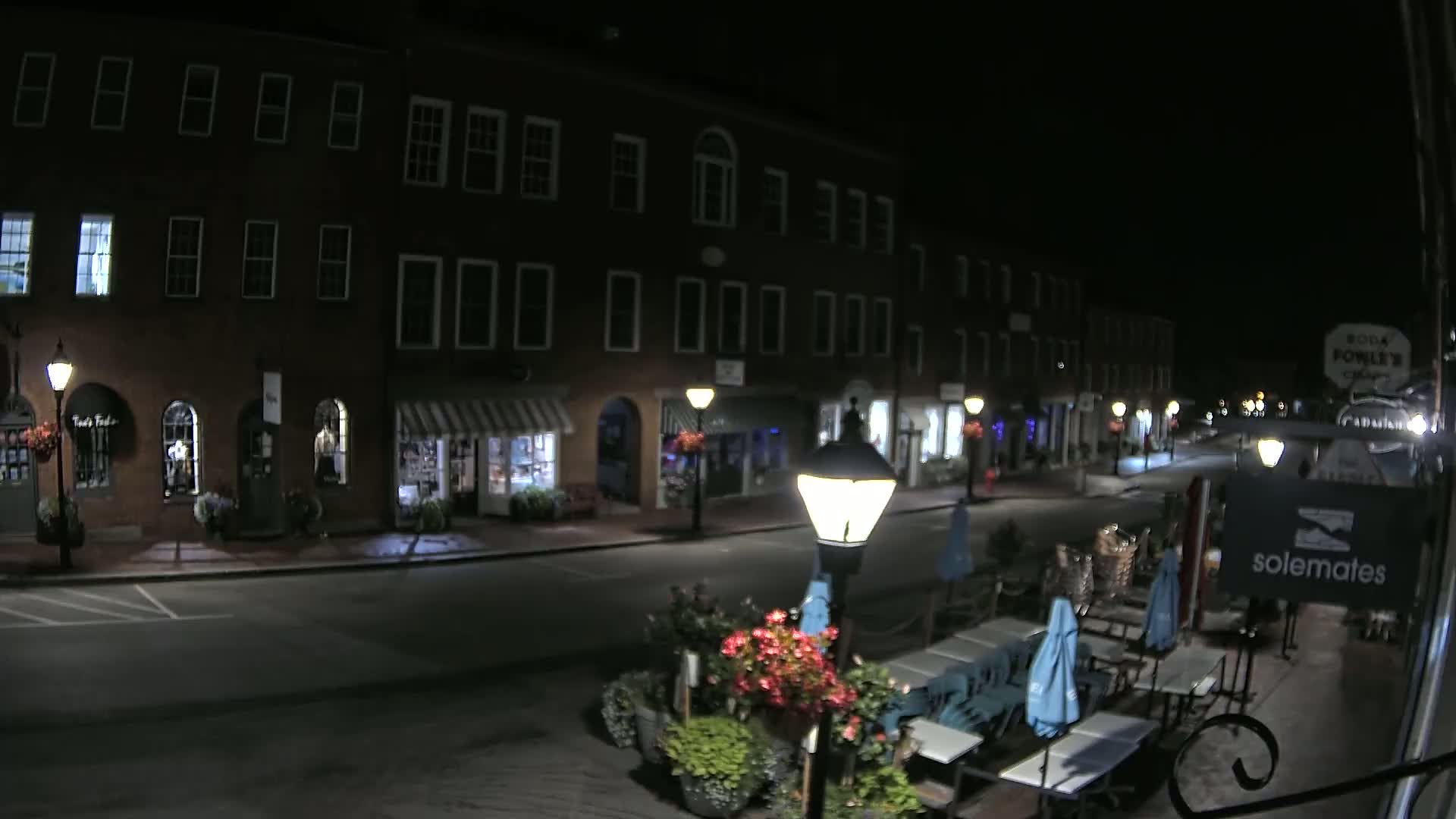 A nighttime street scene shows a row of brick buildings with shops and outdoor seating, illuminated by streetlights under a clear, dark sky.