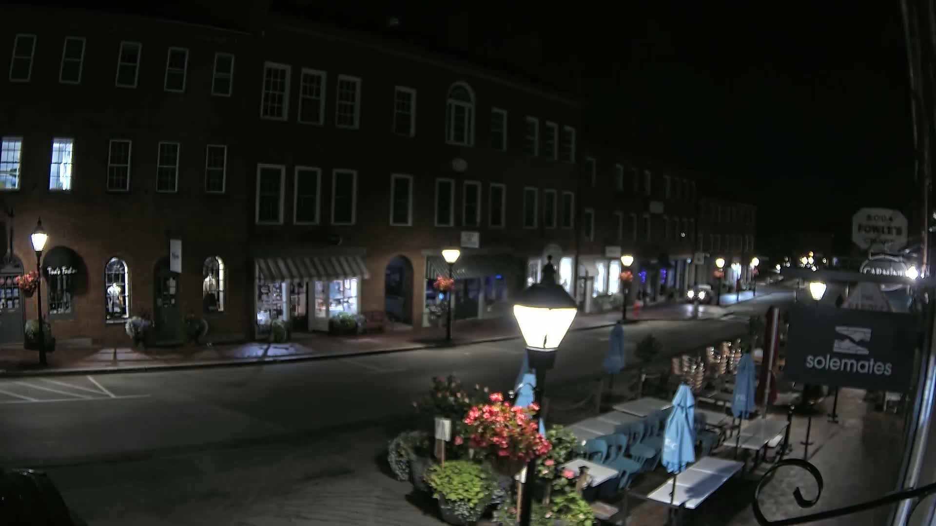 A nighttime street scene shows a row of brick buildings with shops and restaurants, some with outdoor seating, under a clear, dark sky.