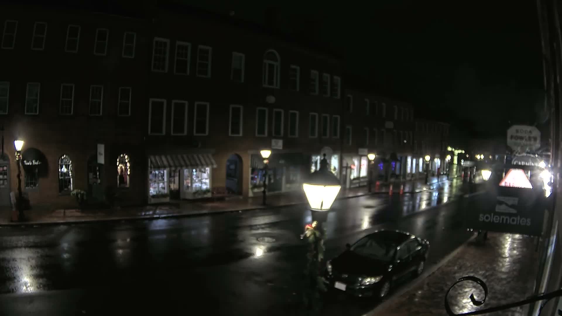 A wet city street is illuminated by numerous streetlights and storefronts at night, with buildings lining the thoroughfare and a car parked near a person.