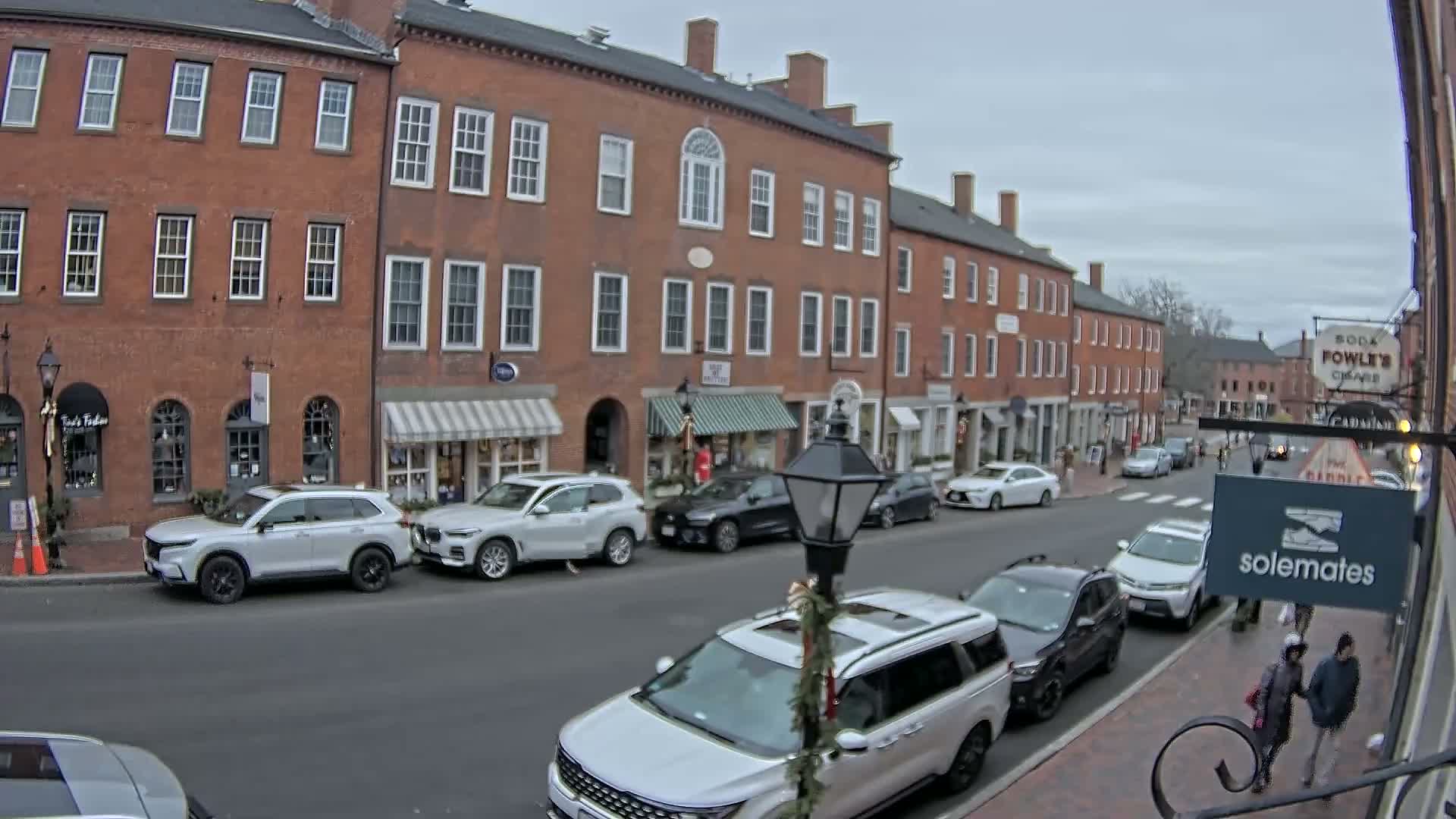 A wet city street is illuminated by numerous streetlights and storefronts at night, with buildings lining the thoroughfare and a car parked near a person.