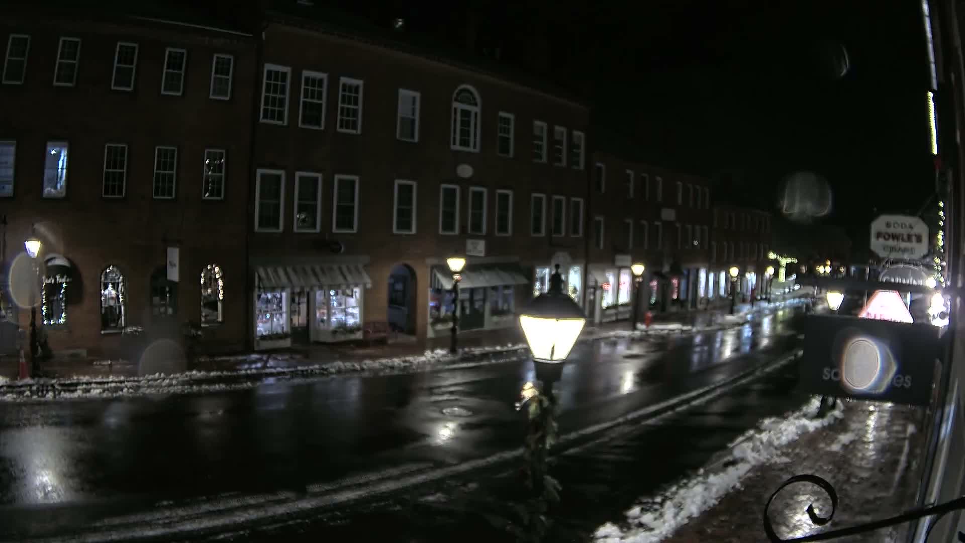 A wintry night scene depicts a quiet street lined with illuminated brick buildings and shops, with patches of snow along the sidewalks and a wet, reflective road.