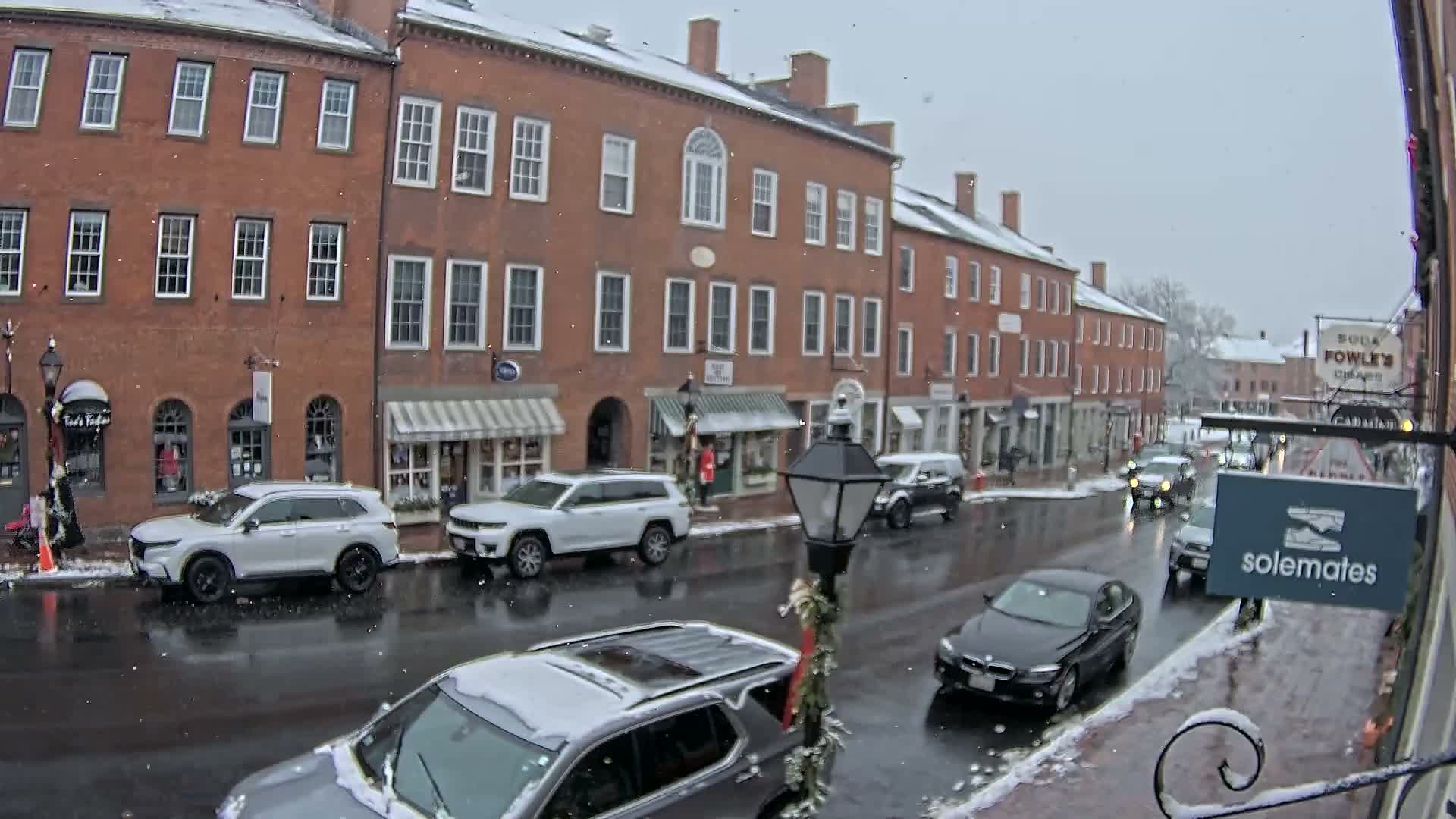 A wintry night scene depicts a quiet street lined with illuminated brick buildings and shops, with patches of snow along the sidewalks and a wet, reflective road.