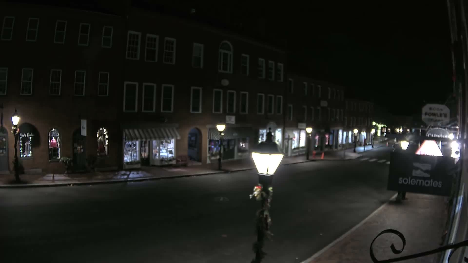 A quiet street at night is illuminated by bright streetlights and the warm glow from multiple shop windows on brick buildings under a clear, dark sky.