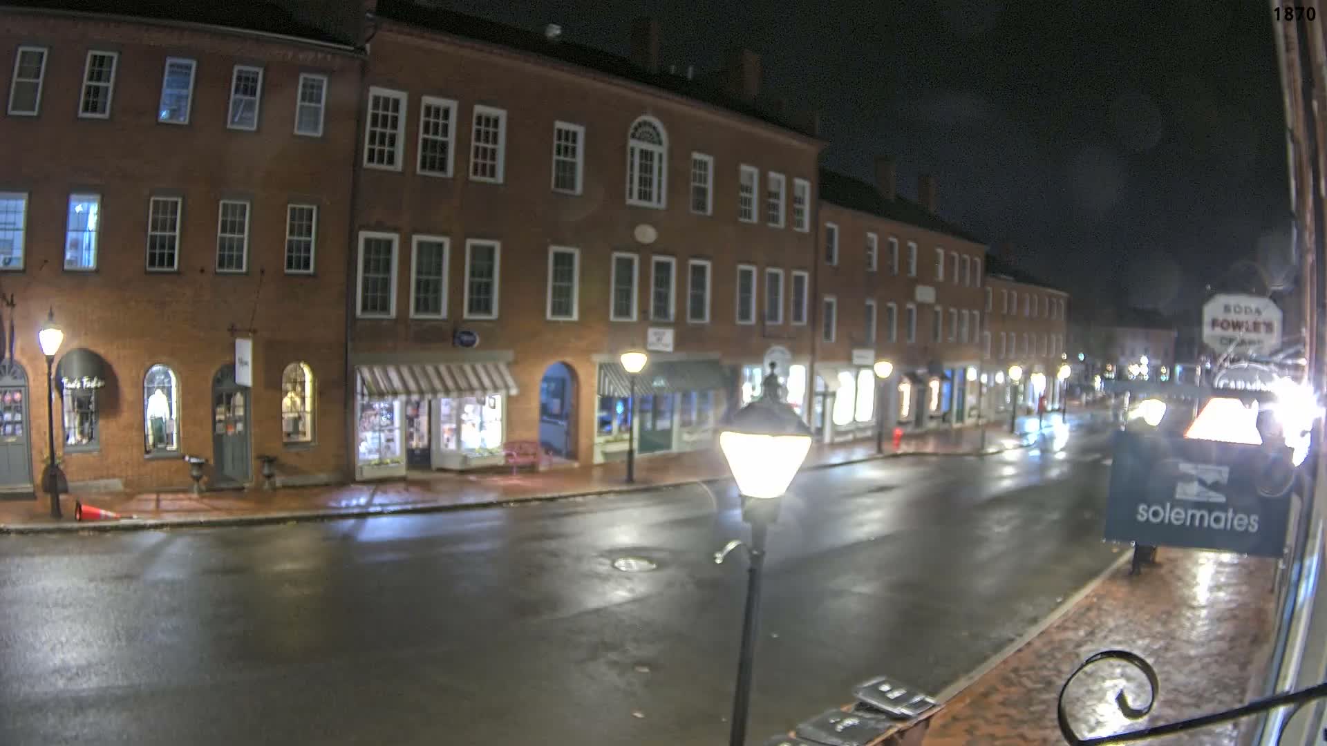 A quiet street at night is illuminated by bright streetlights and the warm glow from multiple shop windows on brick buildings under a clear, dark sky.