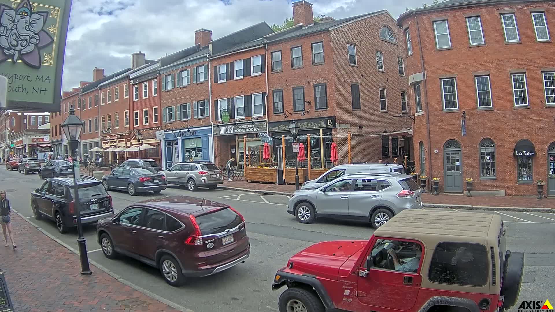 A partly cloudy day on a brick-lined street with parked cars and several pedestrians, featuring multiple restaurants with outdoor seating.