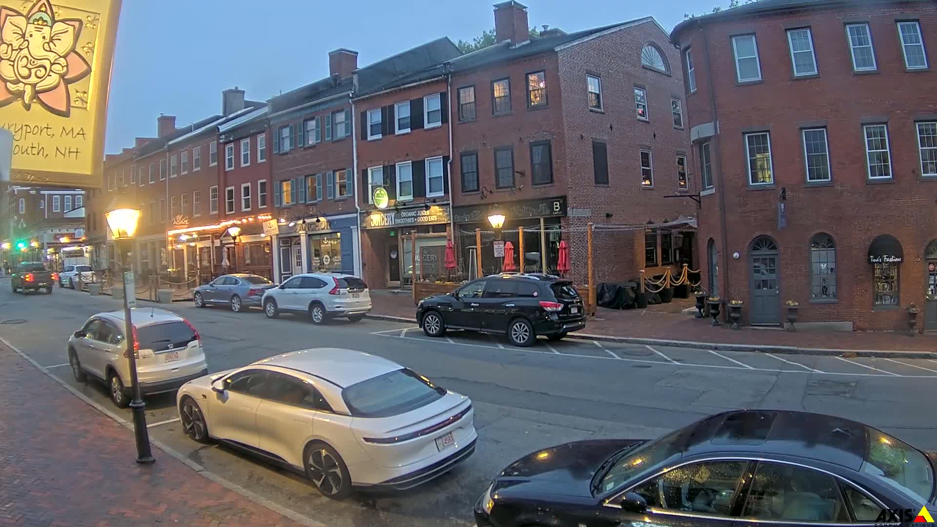 Under a twilight sky, several cars are parked along a street lined with brick buildings, some with outdoor seating areas.