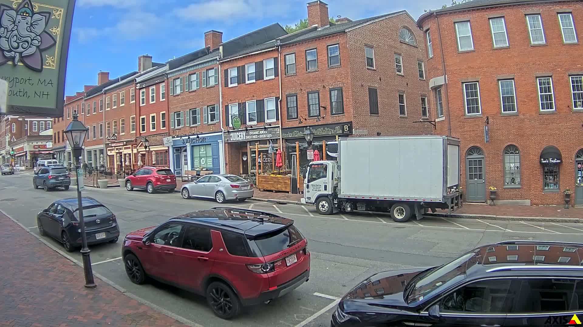A sunny day on a brick-lined street with parked cars and a delivery truck in front of shops and restaurants.