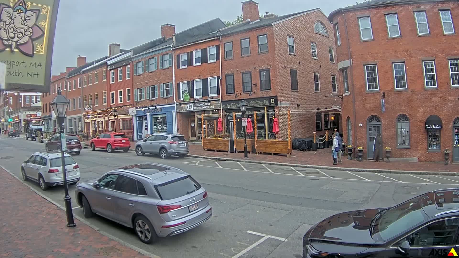 A street scene on an overcast day shows several parked cars in front of brick buildings with various shops and restaurants, including one with outdoor seating.