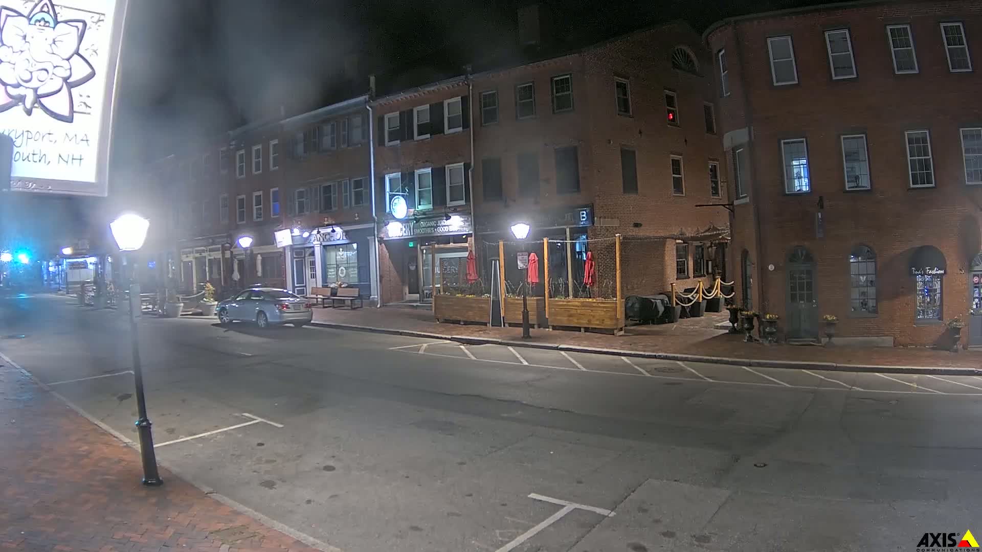 A mostly empty street at night lined with brick buildings, some with outdoor seating, is illuminated by street lamps.