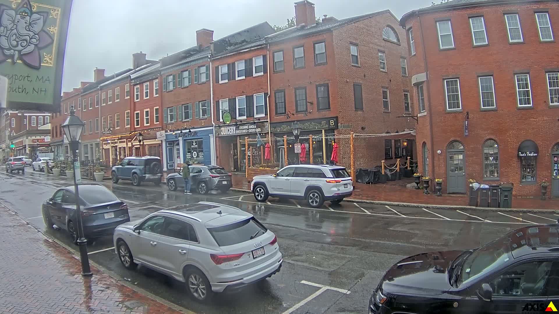 A rainy street scene shows several parked cars in front of brick buildings with various shops and restaurants.