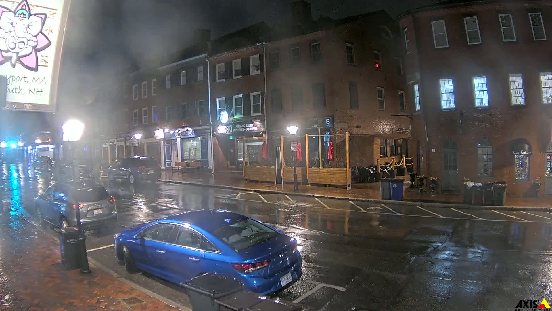 A rain-slicked street at night, lined with brick buildings and parked cars, shows a few businesses with outdoor seating areas.