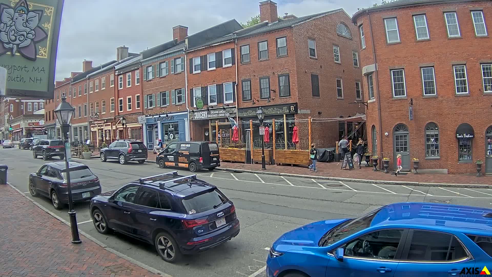On a partly cloudy day, cars are parked along a brick street lined with multi-story brick buildings and small businesses, with several people walking on the sidewalk.