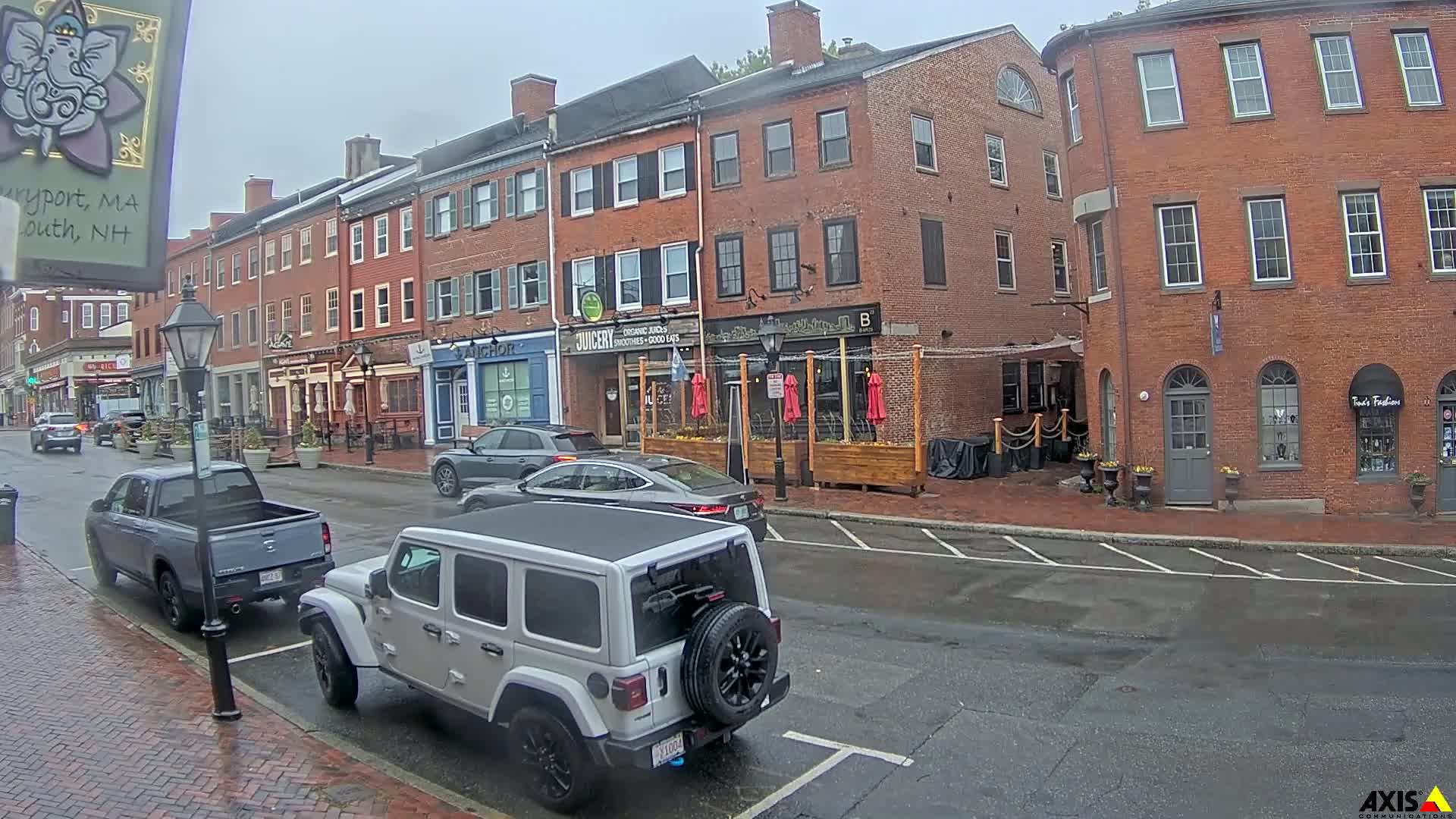 A rainy street scene shows several cars parked and driving past brick buildings with shops and restaurants.