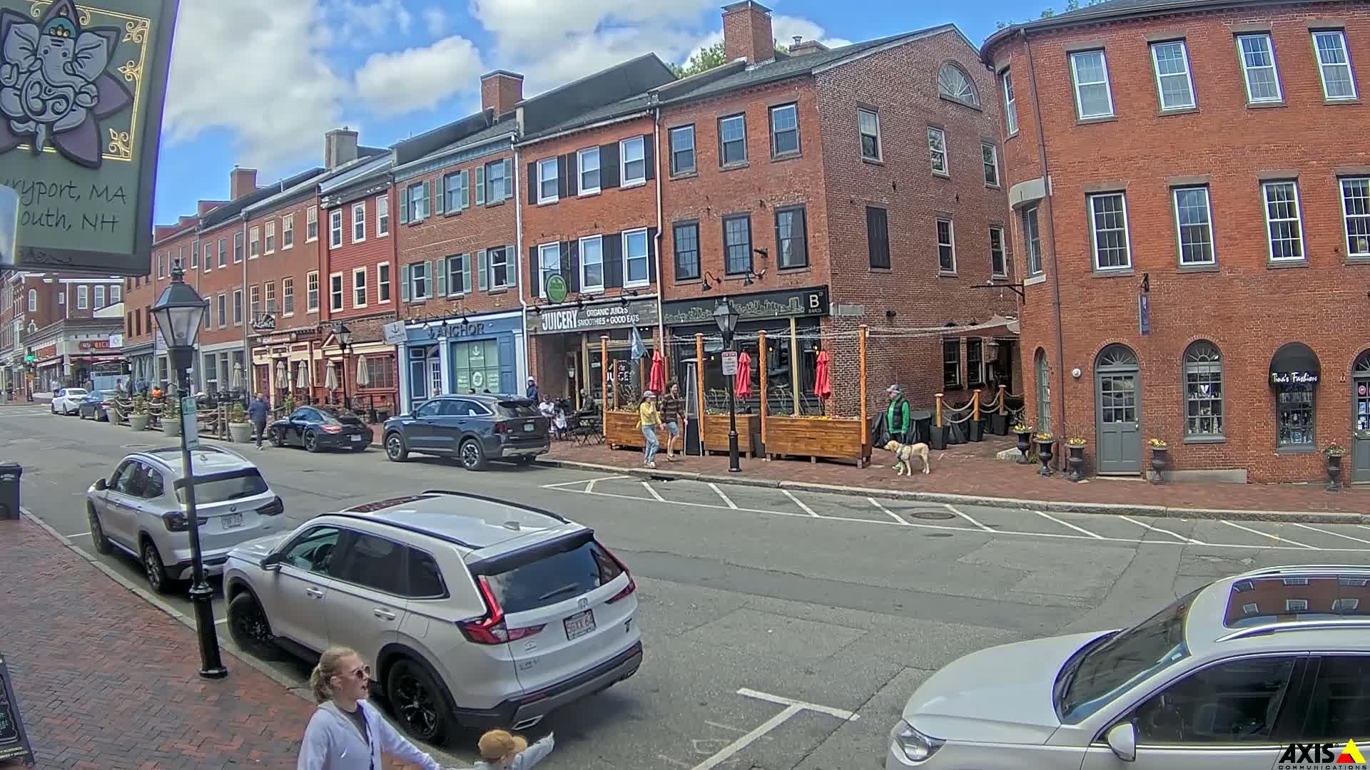 A sunny day on a brick-lined street with parked cars and pedestrians in front of several shops and restaurants, some with outdoor seating.