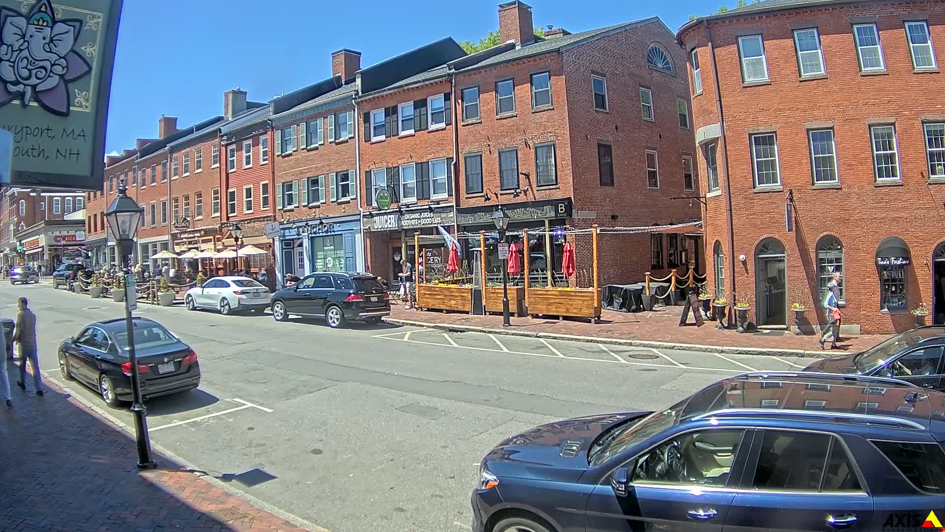 A sunny street scene shows brick buildings with outdoor seating areas in front of restaurants and shops, along with several parked cars and pedestrians.