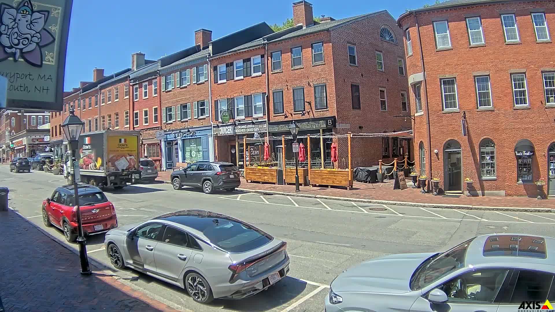 A sunny street scene shows several parked cars in front of brick buildings with outdoor seating areas and businesses.