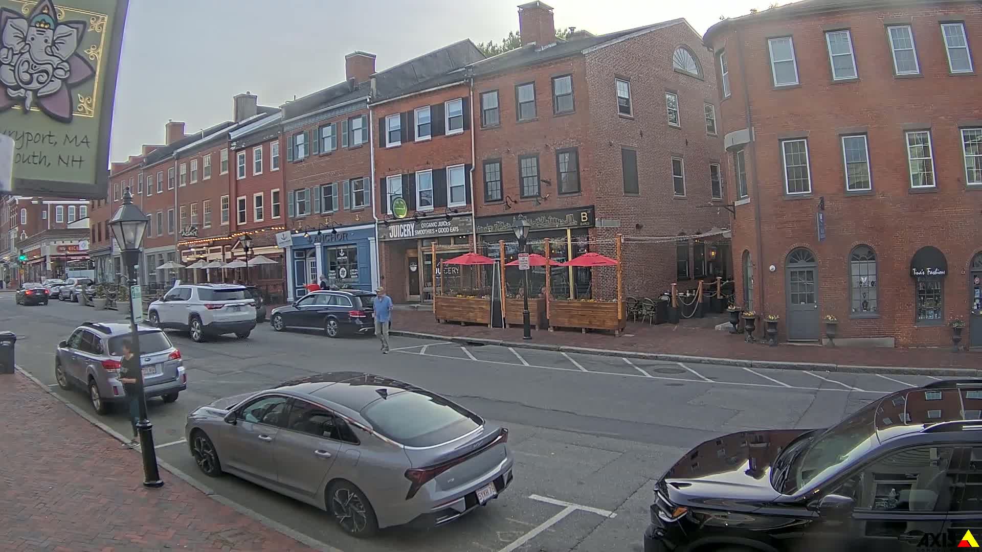A street scene shows several parked cars and a pedestrian in front of brick buildings with outdoor seating areas under red umbrellas on an overcast day.