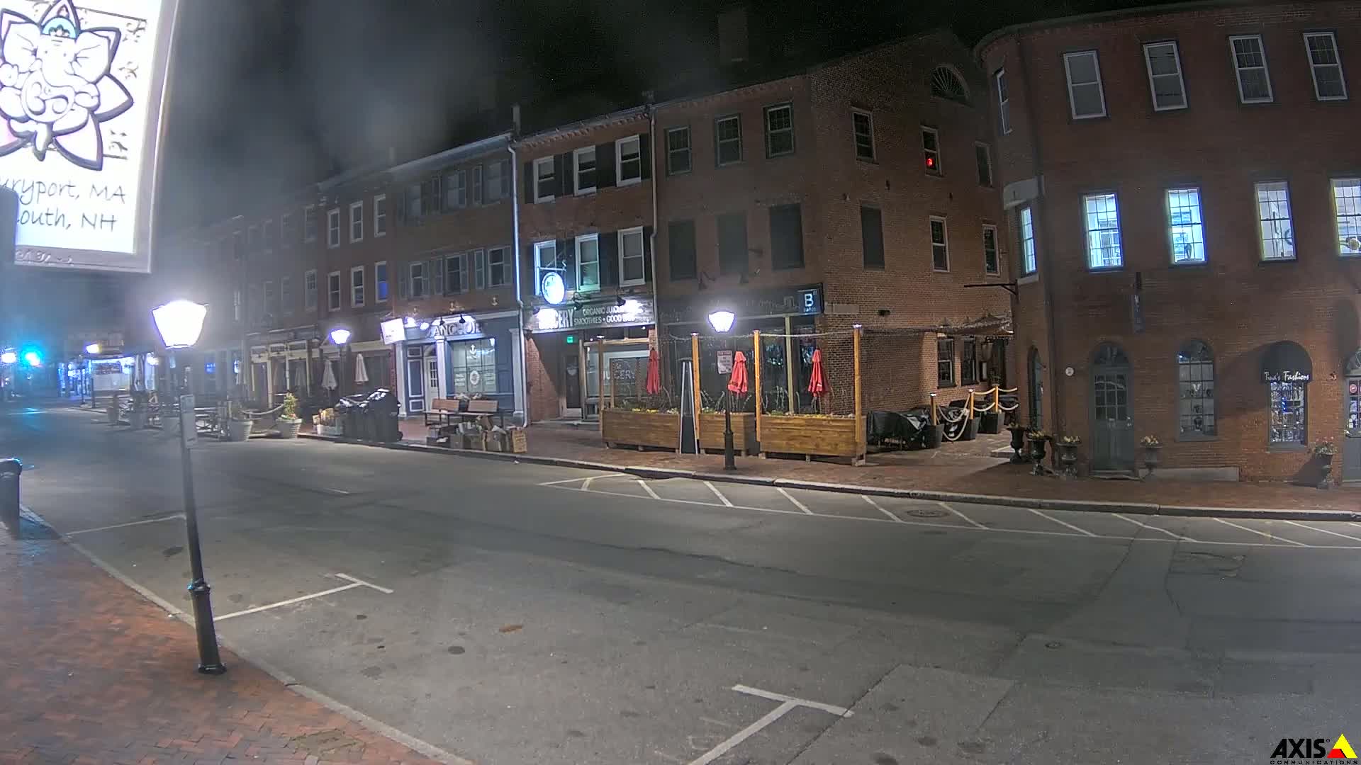 A nighttime street scene shows several brick buildings with outdoor seating areas, illuminated by streetlights under a dark, cloudy sky.