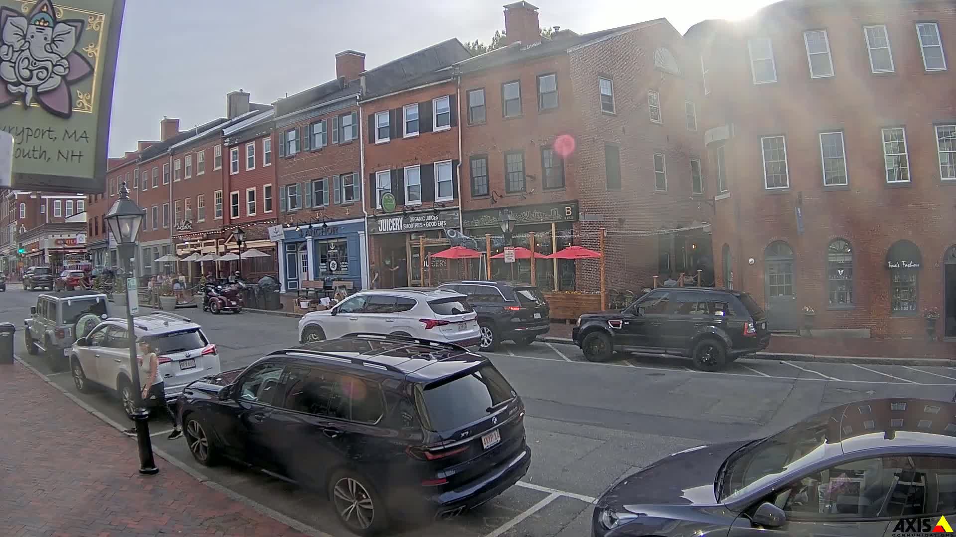 A sunny day on a brick-lined street with parked cars and outdoor seating in front of several shops and restaurants.