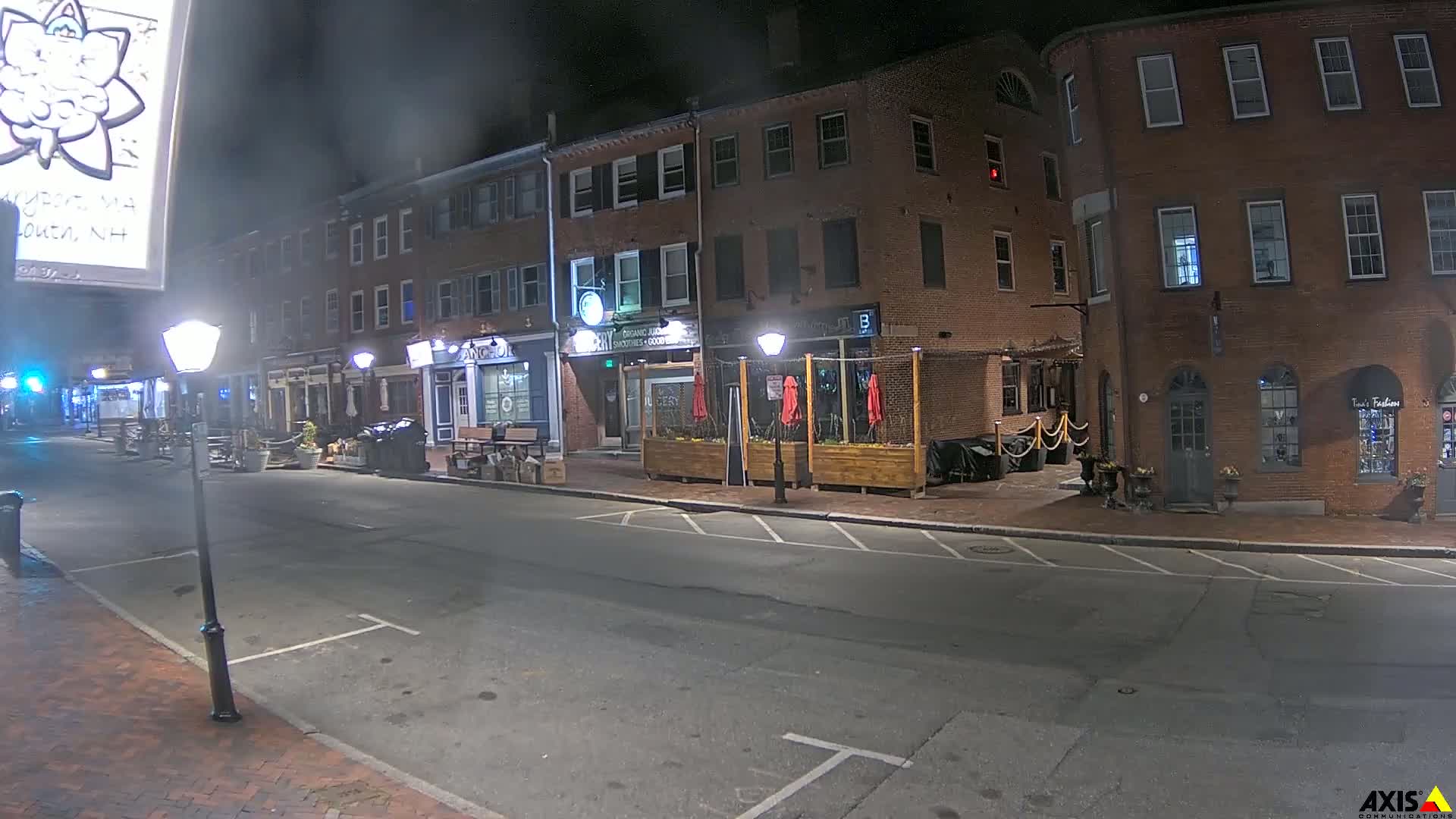 A mostly empty street at night, lined with brick buildings and outdoor seating areas, under hazy, dark conditions.