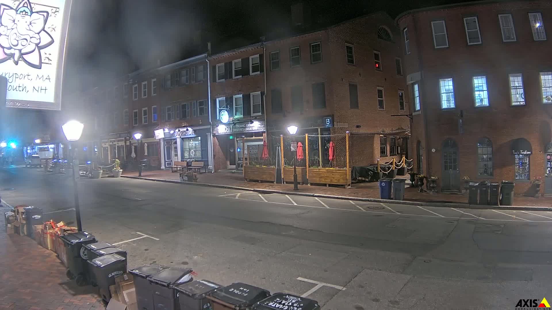A mostly empty street at night lined with brick buildings and outdoor seating areas, under hazy conditions.