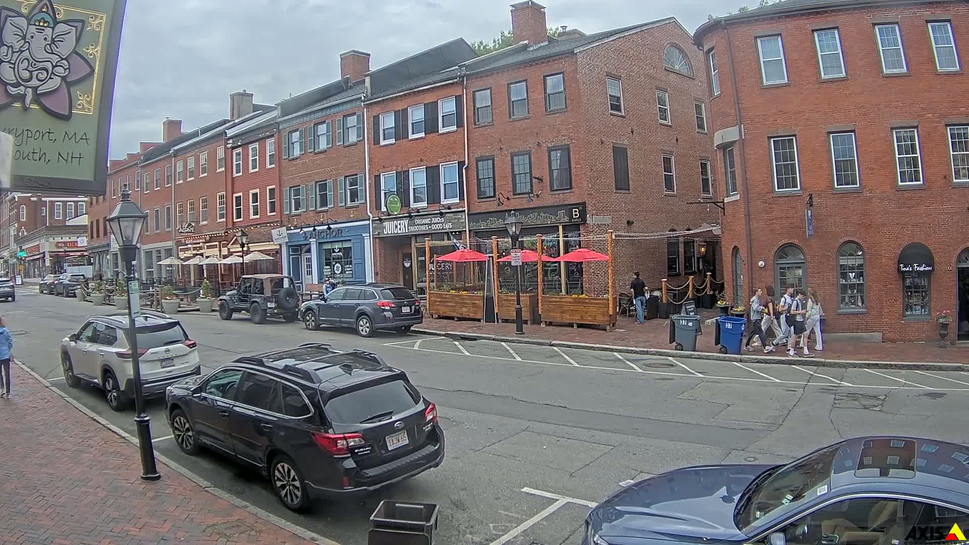 A city street with parked cars and pedestrians on a cloudy day, lined with brick buildings housing restaurants and shops with outdoor seating.