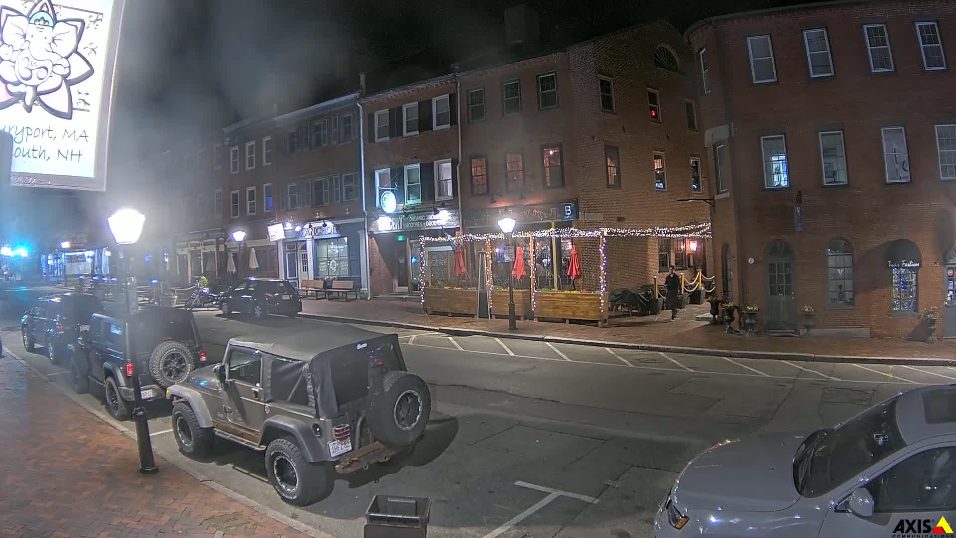A nighttime street scene shows several parked vehicles in front of brick buildings with outdoor seating, string lights, and some businesses illuminated.