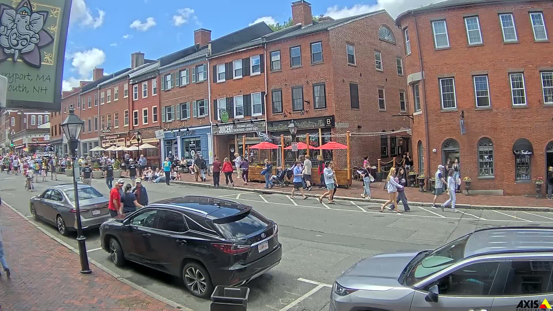 A sunny day on a brick-lined street, crowded with pedestrians and parked cars, in front of several shops and restaurants with outdoor seating.