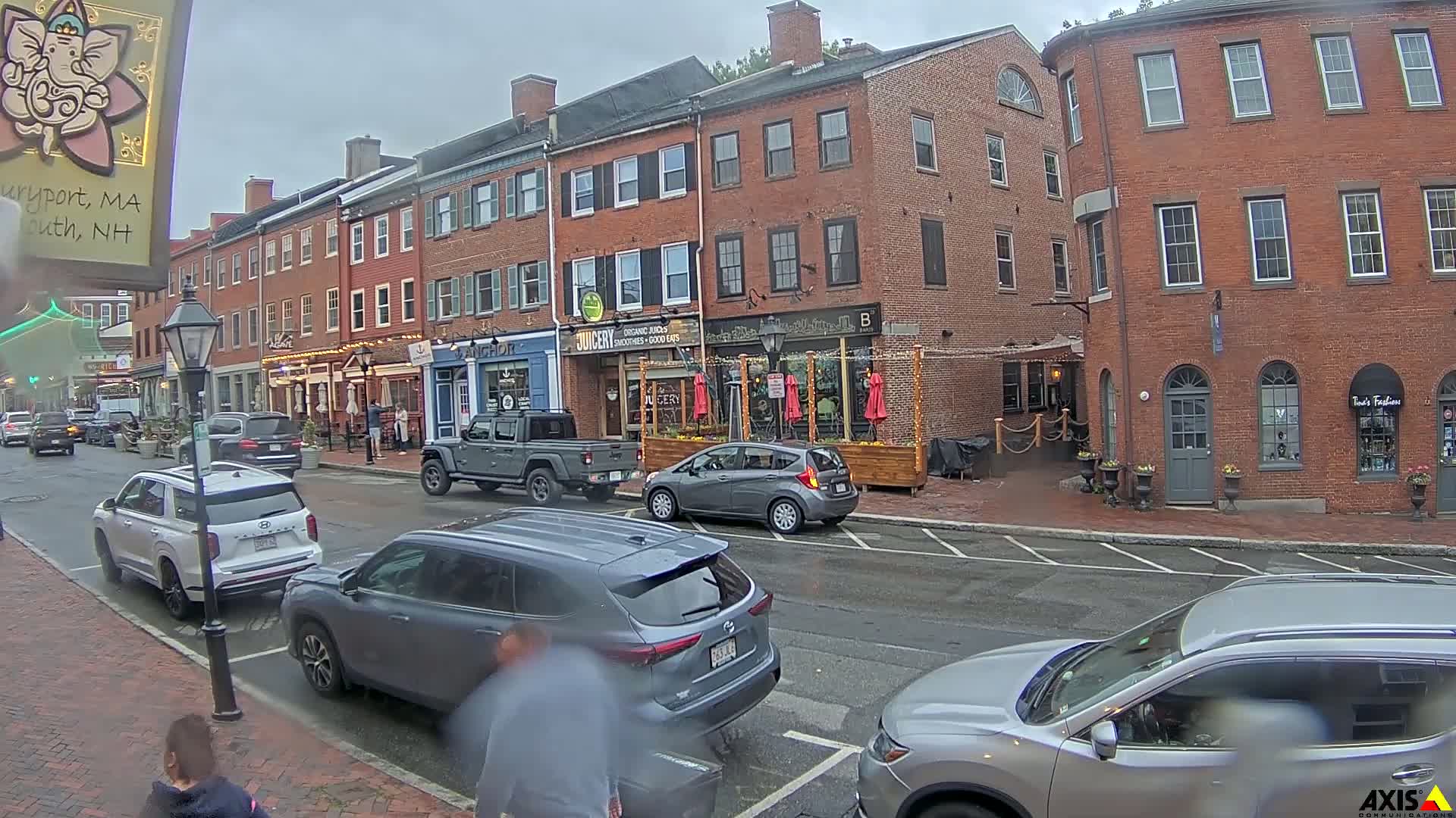 A wet street scene shows several cars parked and driving past brick buildings with outdoor seating areas, under an overcast sky.