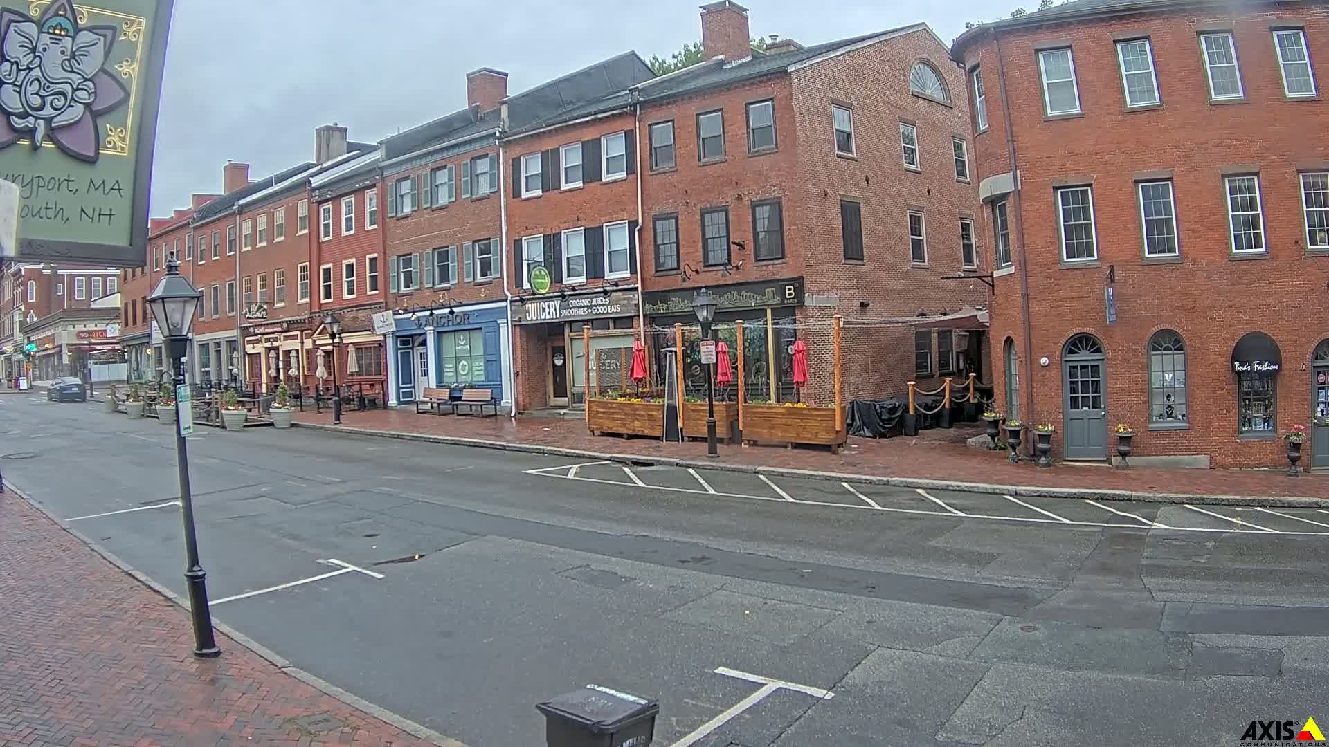 A wet street lined with brick buildings housing various shops and restaurants under an overcast sky.