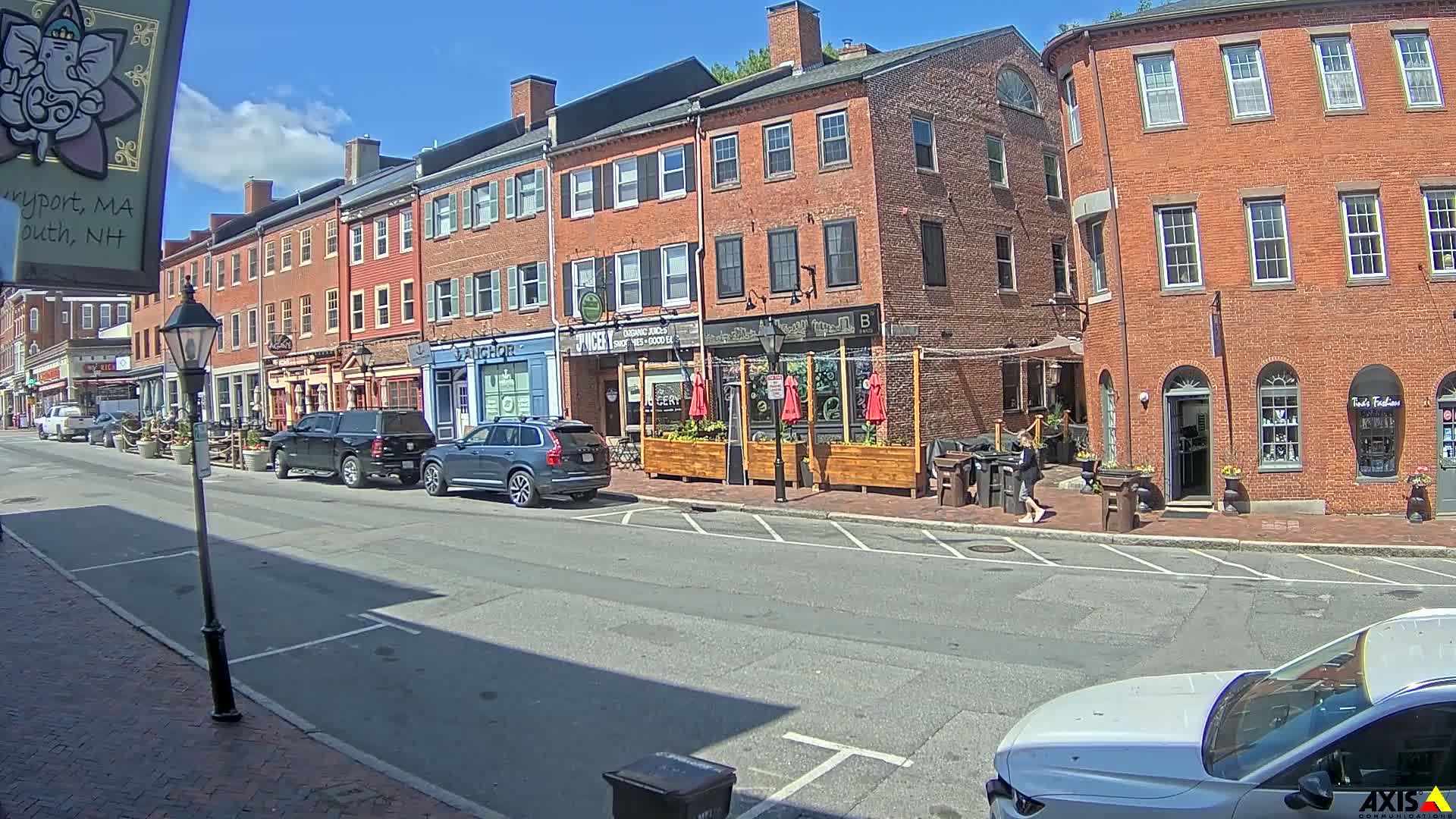 A sunny street scene shows brick buildings with shops and outdoor seating, parked cars, and a person walking.