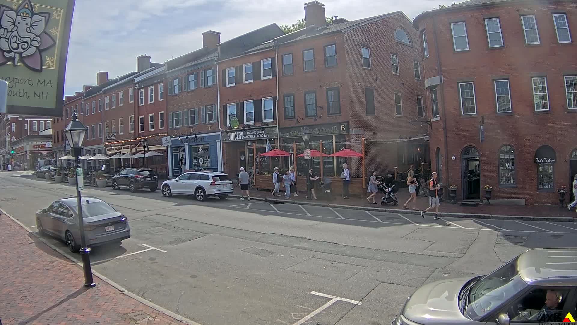 A sunny street scene shows several brick buildings with outdoor seating areas in front of restaurants and shops, along with parked cars and pedestrians.