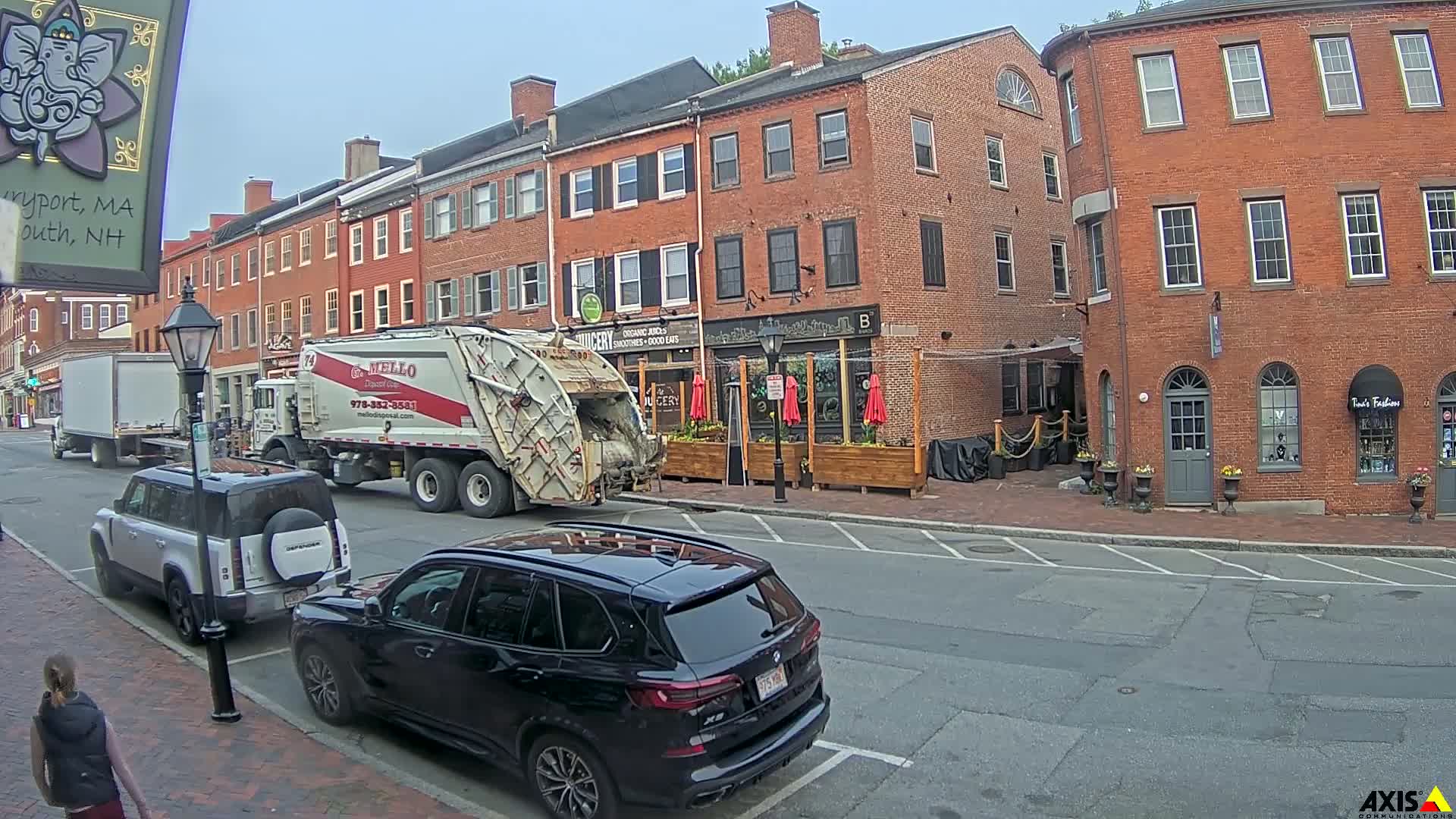 A garbage truck is parked on a city street next to a brick building with outdoor seating, and there are several cars parked along the street on a partly cloudy day.
