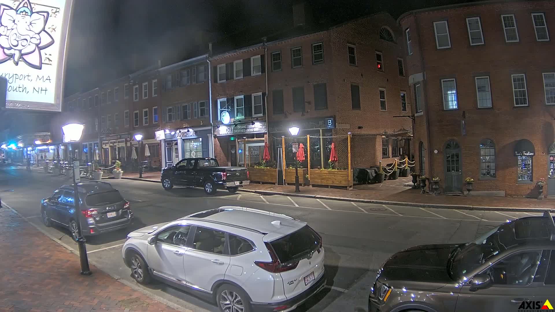 A nighttime street scene shows several parked cars in front of brick buildings with outdoor seating and streetlights.