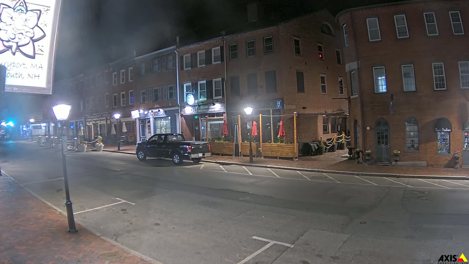 A dark-colored pickup truck is parked on a street at night in front of brick buildings with outdoor seating.