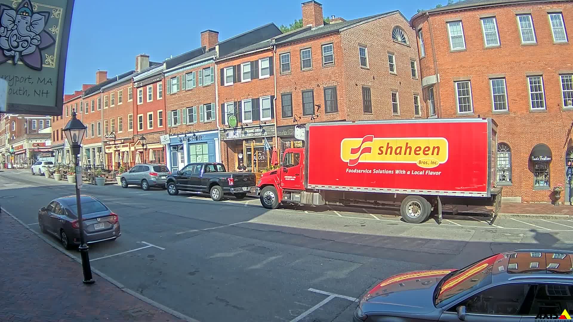 A red Shaheen Bros. Inc. food service truck is parked on a sunny street in front of several brick buildings with outdoor seating.
