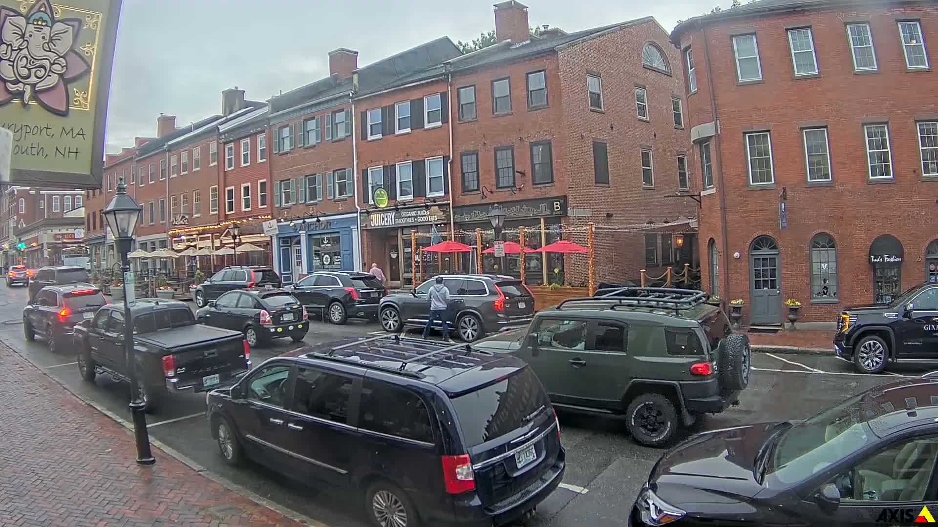 A row of parked cars lines a brick street in front of multi-story brick buildings with outdoor seating areas under an overcast sky.