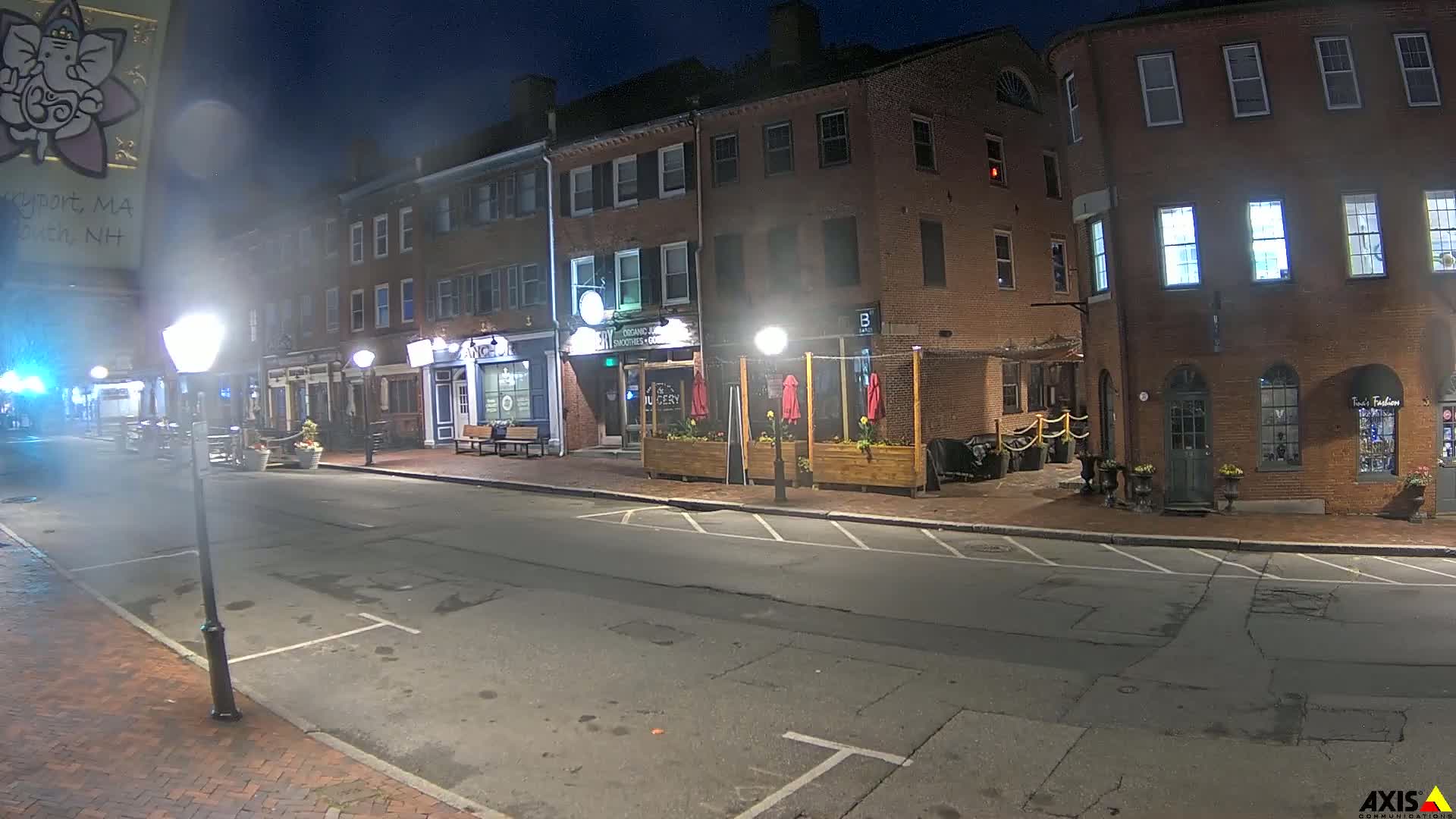 A nighttime street scene shows a row of brick buildings with outdoor seating and lampposts illuminating an empty street.