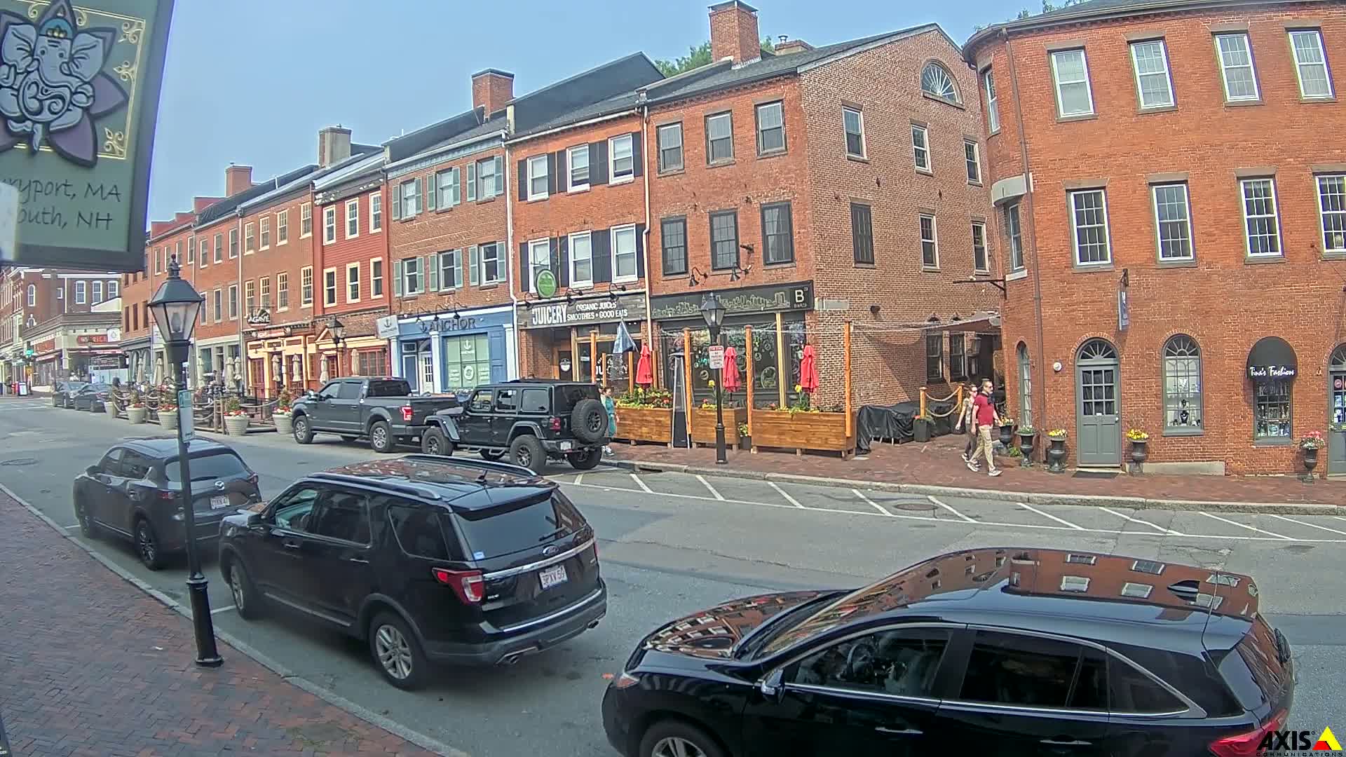 A sunny day on a brick-lined street shows several parked cars in front of shops and restaurants with outdoor seating.